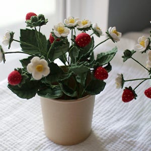 Puede incluir: Una planta de fresas hecha a mano, con flores blancas, hojas verdes y fresas rojas. La planta est&aacute; en una maceta beige y sobre una superficie blanca texturizada. La imagen destaca los detalles artesanales.
