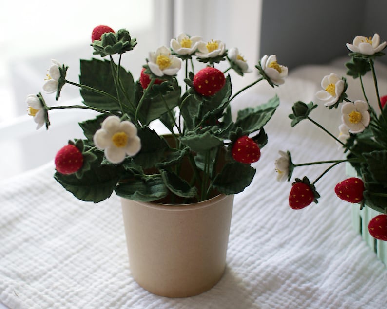 Puede incluir: Una planta de fresas hecha a mano, con flores blancas, hojas verdes y fresas rojas. La planta est&aacute; en una maceta beige y sobre una superficie blanca texturizada. La imagen destaca los detalles artesanales.