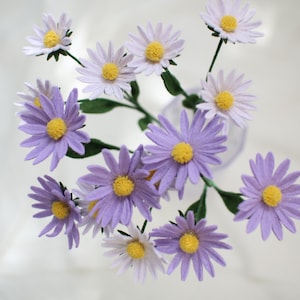 Puede incluir: Primer plano de un ramo de flores de papel hechas a mano. El arreglo presenta flores de color púrpura y blanco con forma de margarita y centros amarillos. Las flores tienen tallos y hojas verdes, creando una estética natural. Las flores están en un jarrón.