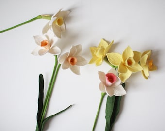 May include: A collection of handmade felt daffodils in various stages of bloom. The flowers feature white, yellow, and pink petals with orange and yellow centers. Green stems and leaves are also visible against a white background.