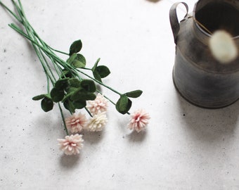 May include: A bouquet of felt flowers with pink and cream-colored blooms and green stems and leaves. The flowers are arranged on a white surface next to a rustic metal pitcher.