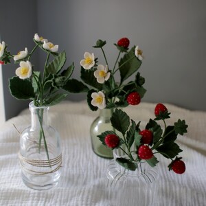 Puede incluir: Tres jarrones de cristal con plantas artificiales de fresas. Las plantas tienen flores blancas con centros amarillos, hojas verdes y fresas rojas con motas blancas. Los jarrones son transparentes y est&aacute;n sobre una tela blanca.