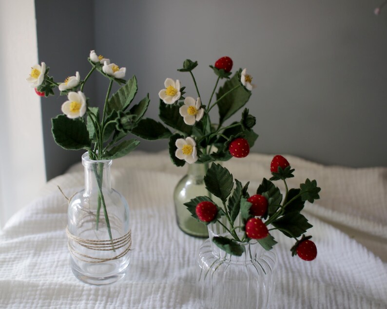 Puede incluir: Tres jarrones de cristal con plantas artificiales de fresas. Las plantas tienen flores blancas con centros amarillos, hojas verdes y fresas rojas con motas blancas. Los jarrones son transparentes y est&aacute;n sobre una tela blanca.