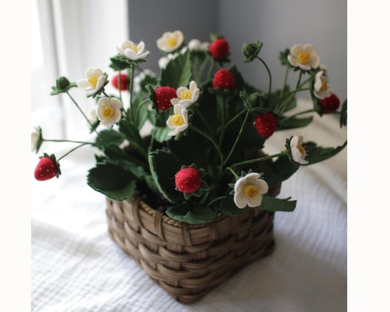 Puede incluir: Una cesta tejida llena de una planta de fresas. La planta presenta hojas verdes, flores blancas con centros amarillos y fresas rojas. La cesta es marr&oacute;n y cuadrada, sobre una superficie blanca.
