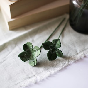 以下が含まれることがあります： 緑色の茎が付いた、2つの緑色のフェルト製の四つ葉のクローバーが、薄緑色のリネン布の上に置かれています。 クローバーには白いアクセントがあり、本の山と濃いガラスの花瓶の近くにあります。