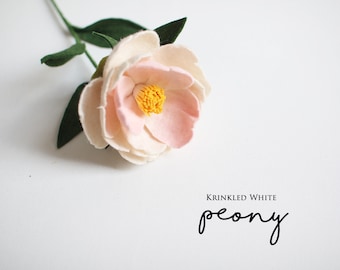 May include: A handmade felt peony flower in shades of cream and light pink, with a yellow center. The flower has green leaves and stem. The text "Krinkled White peony" is below the flower.