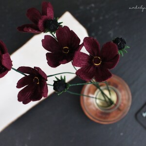 May include: A bouquet of burgundy cosmos flowers with yellow centers in a small glass vase. The flowers have green stems and are arranged on a dark surface. The image includes the text "madebynaomy".