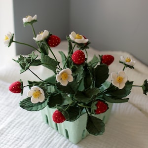 Puede incluir: Un arreglo artesanal de plantas de fresa en una cesta de bayas verde claro. El arreglo presenta fresas rojas con semillas blancas, flores blancas con centros amarillos y hojas verdes. Una pieza decorativa encantadora.