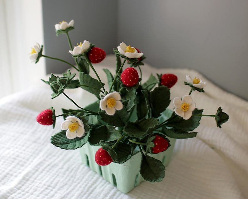 Puede incluir: Un arreglo artesanal de plantas de fresa en una cesta de bayas verde claro. El arreglo presenta fresas rojas con semillas blancas, flores blancas con centros amarillos y hojas verdes. Una pieza decorativa encantadora.