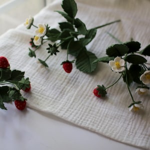 Puede incluir: Varios tallos de fresas artificiales con bayas rojas, flores blancas y hojas verdes est&aacute;n dispuestos sobre una superficie blanca y texturizada. Las fresas tienen un aspecto texturizado y las flores tienen centros amarillos.