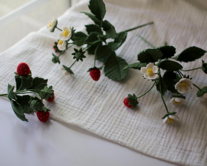 Puede incluir: Varios tallos de fresas artificiales con bayas rojas, flores blancas y hojas verdes est&aacute;n dispuestos sobre una superficie blanca y texturizada. Las fresas tienen un aspecto texturizado y las flores tienen centros amarillos.