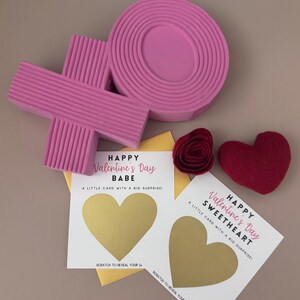 May include: A Valentine's Day gift set featuring a pink 'XOXO' design, a felt heart, a felt rose, and two scratch-off cards with gold hearts. The cards read "Happy Valentine's Day" and "Babe" or "Sweetheart".