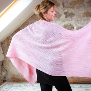 May include: A pink knitted shawl with a delicate open weave pattern. The shawl is draped over a person's shoulders and held in their hands.