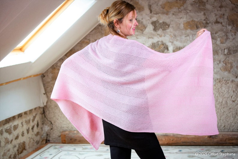 May include: A pink knitted shawl with a delicate open weave pattern. The shawl is draped over a person's shoulders and held in their hands.