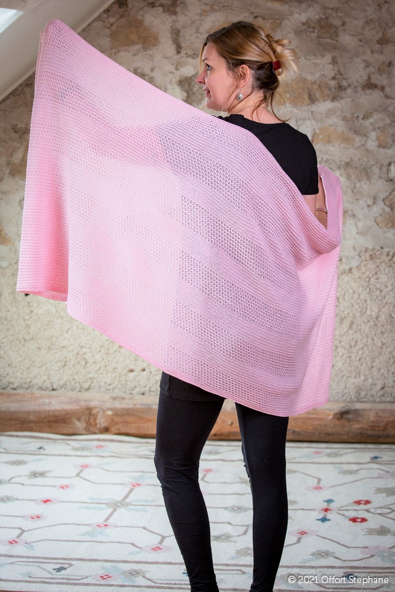May include: A pink knitted shawl with a delicate openwork pattern. The shawl is draped over a person's shoulders and is held in place by their hand.