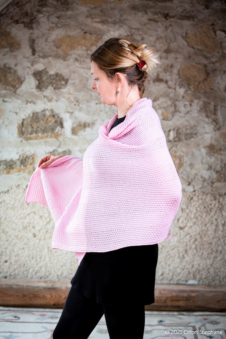 May include: A woman wearing a pink knitted shawl over a black top and black pants. The shawl is a light pink color and has a delicate knit pattern.