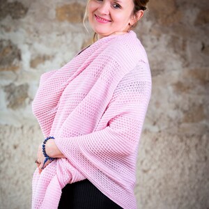 May include: A woman wearing a pink knitted cardigan over a black dress. She is standing in front of a stone wall.
