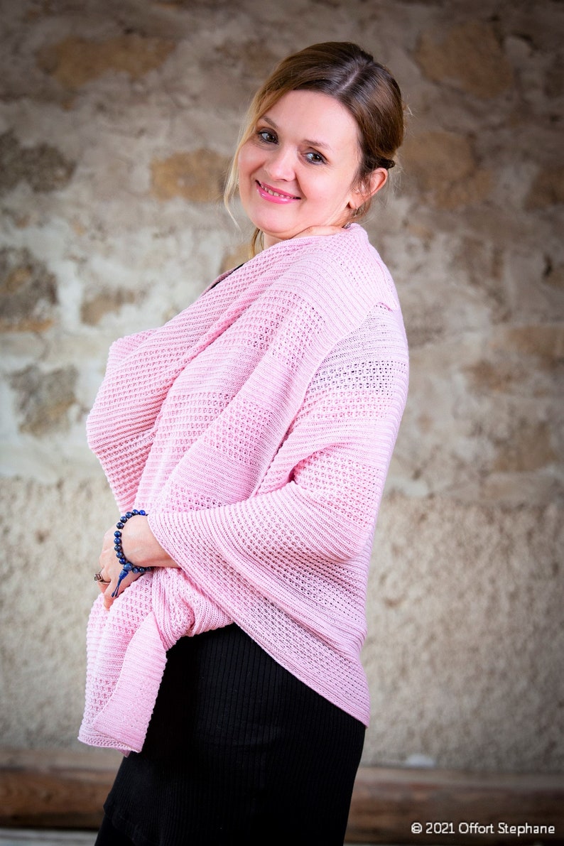 May include: A woman wearing a pink knitted cardigan over a black dress. She is standing in front of a stone wall.