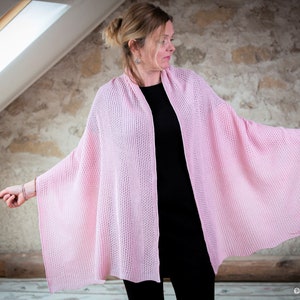 May include: A pink knitted shawl draped over a person's shoulders. The shawl is made of a delicate, open-weave fabric and has a soft, flowing drape.