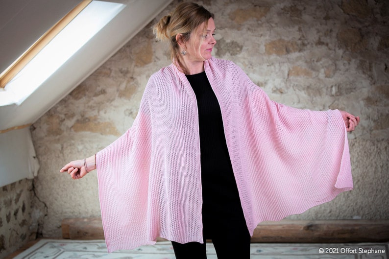 May include: A pink knitted shawl draped over a person's shoulders. The shawl is made of a delicate, open-weave fabric and has a soft, flowing drape.
