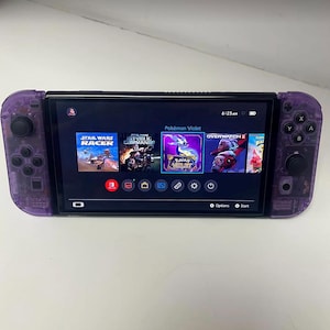 May include: A purple Nintendo Switch console with a black screen displaying the game Pokemon Violet. The console has a purple and black controller on each side. The screen shows the game's title, a purple and pink dragon, and the words "Pokemon Violet".