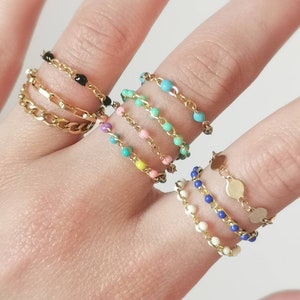 Puede incluir: Un conjunto de anillos de cadena de oro con cuentas de colores. Los anillos están apilados en un dedo, con el anillo superior con cuentas negras, seguido de un anillo turquesa, un anillo rosa, un anillo amarillo, un anillo azul y un anillo blanco. El anillo inferior es una cadena de oro con pequeños abalorios redondos de oro.