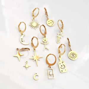 Puede incluir: Un conjunto de 10 pendientes de aro dorados con diferentes colgantes, incluyendo un sol, una luna, estrellas, un muñeco de palo, una botella y un ojo. Los colgantes son todos de metal dorado y están unidos a los aros con un pequeño lazo.