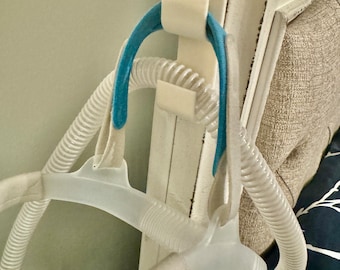 May include: A white and blue CPAP mask with a clear hose is hanging on a white hook attached to a headboard. The hook is secured with adhesive.
