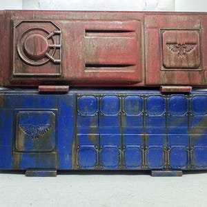 May include: Two-piece, weathered-look storage containers. The top container is red with a circular design and a rectangular emblem. The bottom container is blue with a grid of square details and an emblem. Both have a distressed, aged appearance.