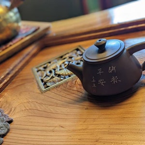 Op de afbeelding: Een bruine keramische theepot met een gebogen handvat en tuit, met Chinese karakters. De theepot staat op een houten oppervlak, naast een goudkleurige theepot en een decoratief vierkant met gesneden details.