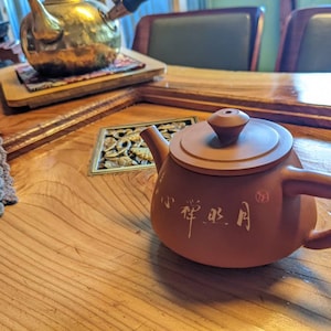 Könnte beinhalten: Eine kleine, rotbraune Keramik-Teekanne mit rundem Körper, gebogenem Henkel und Deckel. Die Teekanne hat chinesische Schriftzeichen auf der Seite. Im Hintergrund steht eine goldfarbene Teekanne auf einem Holztablett.