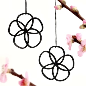 Könnte beinhalten: Zwei schwarze Metallornamente in Blumenform, die an Schnüren vor einem weißen Hintergrund hängen. Die Ornamente bestehen aus überlappenden Kreisen, die eine Blumenform bilden. Der Hintergrund zeigt einen Zweig mit rosa Blüten.