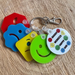 May include: A keyring with colourful acrylic charms depicting different emotions. The charms include red, blue, yellow, and green shapes with faces, and a white disc with the words "Angry", "Worried", "Happy", and "Sad".