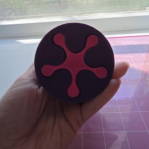 May include: A round, dark purple object with a five-pointed, red design in the center. The object is held in a hand, and the background includes a window and a pink cutting mat.