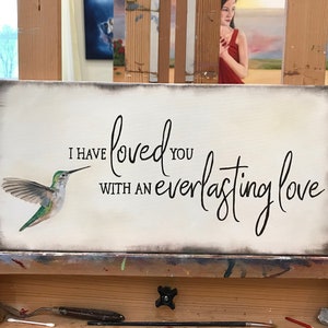May include: A rectangular canvas sign with a distressed grey border on a cream background. It features a painted green and white hummingbird on the left and the black cursive text, I HAVE loved YOU WITH AN everlasting love.