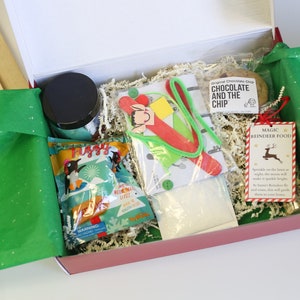 May include: A Christmas-themed gift box filled with various items. Includes a jar with a black lid, a bag of treats, a cookie, and a tag that reads "Magic Reindeer Food". The box is lined with green tissue paper.