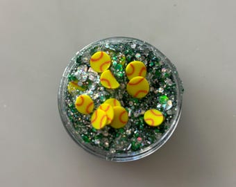 May include: A clear, round container filled with green and silver glitter and small yellow softball-shaped embellishments with red stitching. The container is on a white surface.
