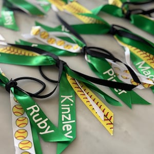 May include: Green and white softball hair ties with names printed on them. The ribbons have softball graphics and are tied with black elastic bands. The background is a light-colored surface.