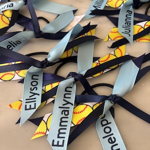 May include: Softball-themed hair ties with navy blue and light blue ribbons. Each tie features a softball pattern and a personalized name printed on a light blue ribbon. The ties are knotted together.