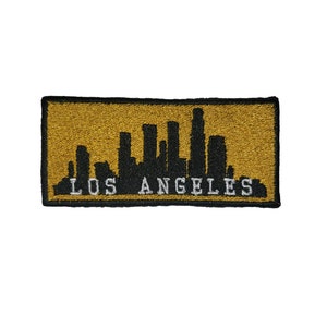 Könnte beinhalten: Ein gold gestickter Aufnäher mit einer schwarzen Silhouette der Skyline von Los Angeles und dem Text "LOS ANGELES" in Weiß.