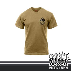 May include: Tan short-sleeve t-shirt with a black USN anchor design on the upper left chest. The shirt has a crew neck and is made of a soft material. The bottom of the image has the text "STEEL beach DESIGN + VINYL".