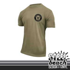 May include: Olive green short-sleeved t-shirt featuring a black circular emblem with the text "UNITED STATES NAVY VETERAN". The shirt has a crew neck and a small patch on the left sleeve. The bottom right corner has the text "STEEL beach DESIGN + VINYL".