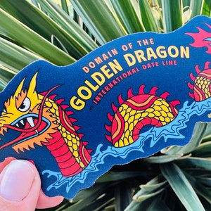 Peut inclure: Un autocollant bleu avec un dragon jaune, rouge et orange avec le texte "Domain of the Golden Dragon - International Date Line" et le nom de profil sur les réseaux sociaux "@steelbeachdesigns".