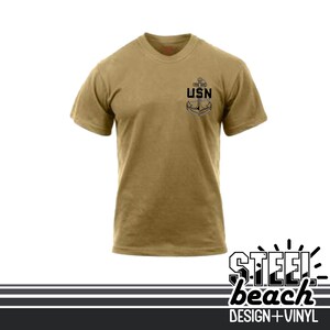 May include: A tan t-shirt with a crew neck and short sleeves. The shirt features a black and gray anchor design with the letters "USN" on the upper left chest. The bottom right corner of the image has the text "Steel Beach Design + Vinyl".