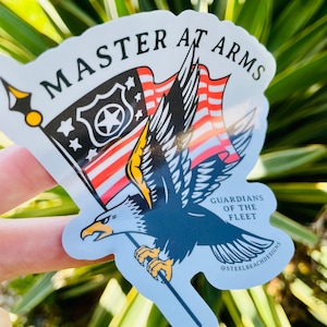 Puede incluir: Una pegatina blanca con una ilustración en blanco y negro de un águila sosteniendo una bandera con el texto "MASTER AT ARMS" y "GUARDIANS OF THE FLEET" con el texto "@STEELBEACHDESIGNS" en la parte inferior.
