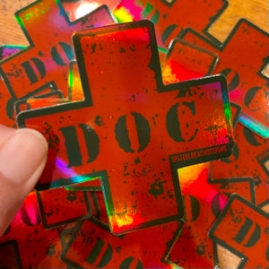 Puede incluir: Una pegatina holográfica roja con una cruz blanca y la palabra "DOC" en letras negras. La pegatina tiene un aspecto desgastado y el texto "@STEELBEACHDESIGNS" está impreso en la esquina inferior derecha.