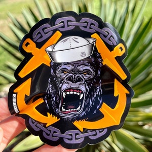 May include: A black and yellow sticker with a gorilla wearing a white sailor hat and a chain around its head. The gorilla is roaring and has an anchor on each side of its head. The sticker has the text "STEELBEARD" on the bottom left corner.