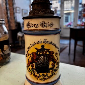 以下が含まれることがあります： ダークメタルの蓋が付いた、ヴィンテージのセラミック製ビールジョッキ。ジョッキはクリーム色で、青と茶色のアクセントが付いています。「Georg Reis」という名前と、ライオンと紋章のエンブレムが特徴です。