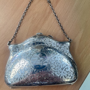 May include: A silver-colored, ornate purse with a chain handle. The purse features intricate floral engravings across its surface. Text on the purse reads "Sister to From U.M.C. High St Longton For 21 Years Valuable Service July 1921."