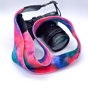 May include: A black camera with a lens and a colourful tie-dye camera strap. The strap features a vibrant mix of pink, blue, and green hues. The camera is positioned on a white background.